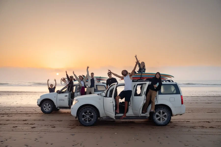 Group of guests posing with raised arms on white vehicles with surfboards during sunset at Aourir beach.