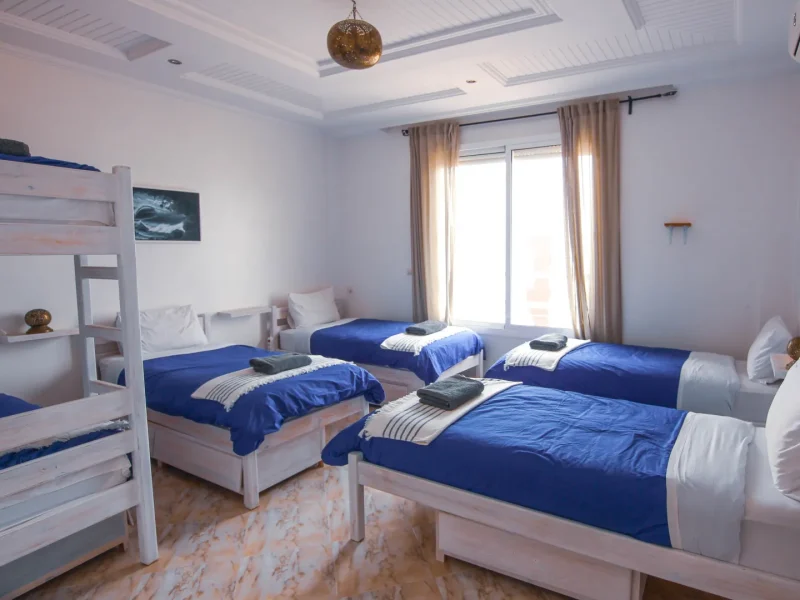 Male shared dorm room at Sunshine Surf Morocco in Taghazout with four single beds with blue duvets, white woodwork, and contemporary furnishings.