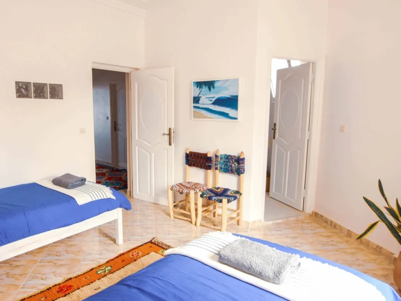 Twin ensuite room at Sunshine Surf Morocco in Taghazout with two blue beds, white walls, and tiled flooring.