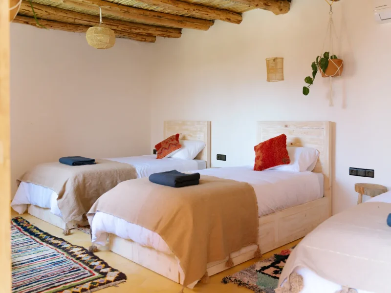 Triple bedroom at Sunshine Surf Morocco in Taghazout features twin wooden beds with red accent pillows, white linens, and exposed timber ceiling beams.