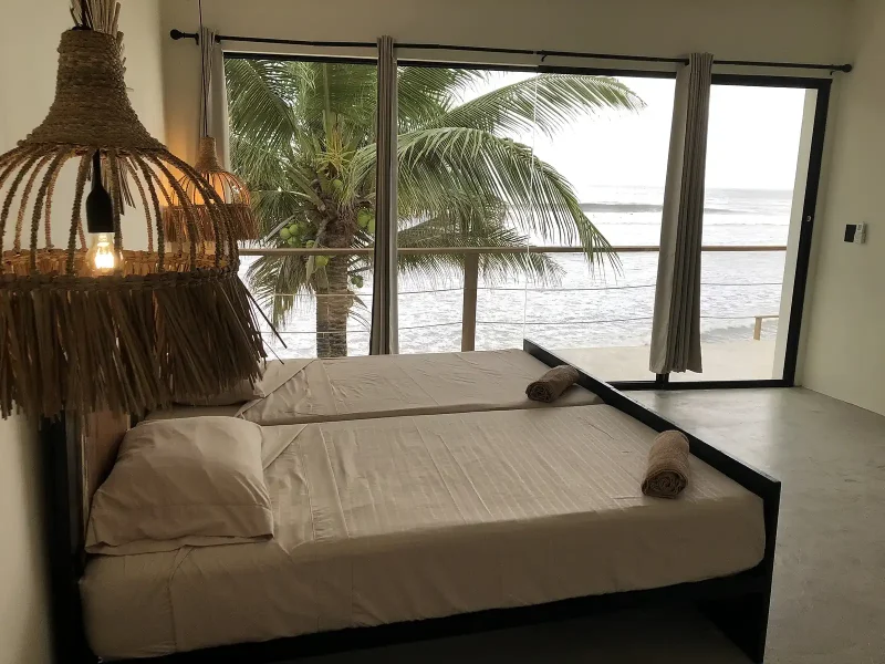 Doppelzimmer mit weißer Bettwäsche und Meerblick durch bodentiefe Fenster zum Strand von El Sunzal, mit sichtbaren Palmen draußen.
