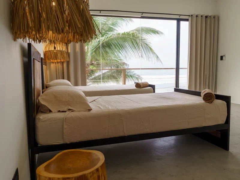 Geräumiges Schlafzimmer im Laola Surf Camp El Sunzal mit großem Bett, bodentiefen Fenstern mit Blick auf Ozean und Palmen sowie Rattan-Hängebeleuchtung.