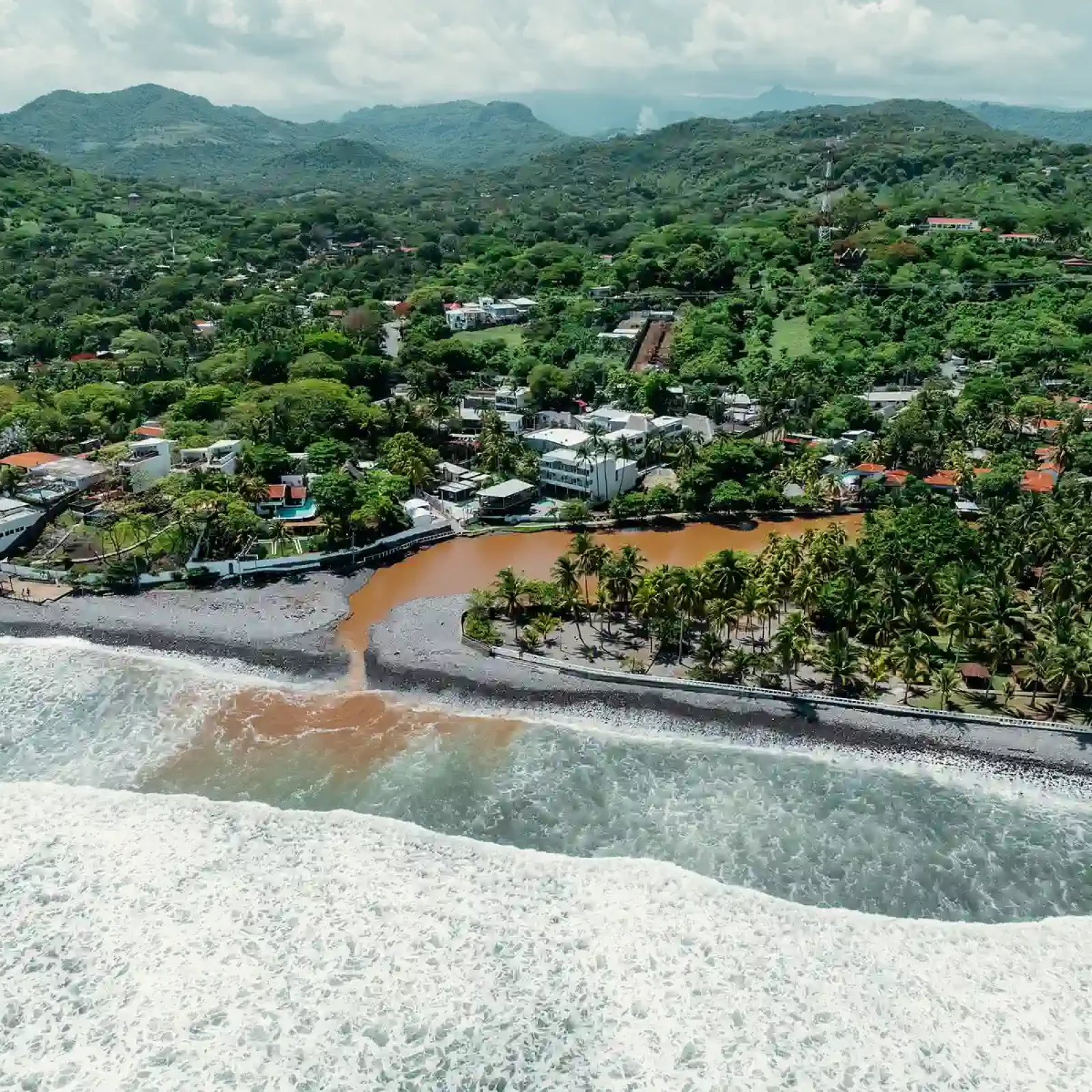 Vue aérienne de l'établissement côtier El Sunzal montrant l'embouchure de la rivière brune rencontrant la plage de sable blanc avec végétation verte et montagnes en arrière-plan.