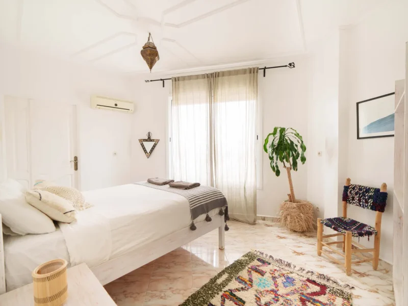 Light-filled bedroom with double bed, white walls, sheer curtains, potted plant and Moroccan decor at Sunshine Surf Morocco.