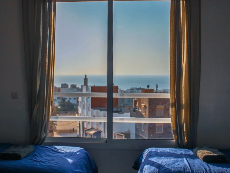 Female shared dorm room with two blue beds facing windows overlooking Taghazout coastal town and Atlantic Ocean.