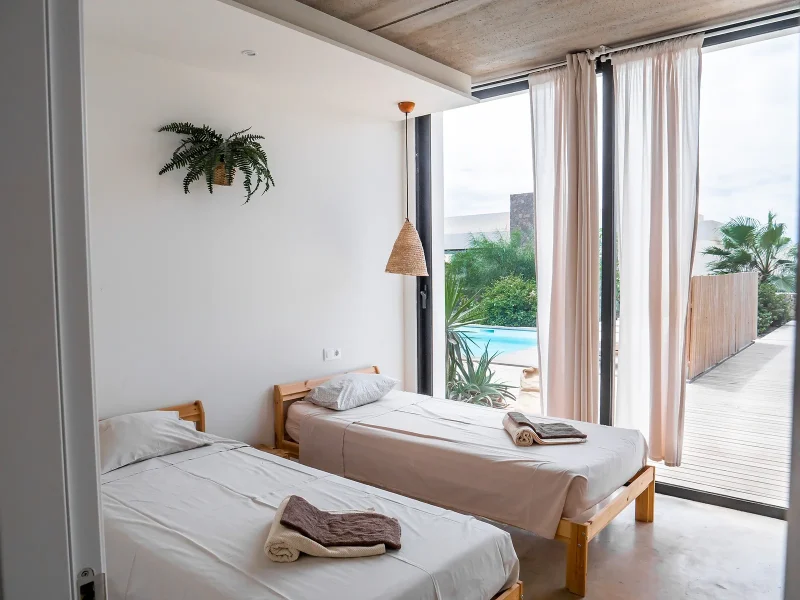 Zwillingsschlafzimmer mit Holzbettrahmen, weißer Bettwäsche und offenem Terrassen-Zugang mit Blick auf Pool und tropische Gärten im Laola Surf Camp.