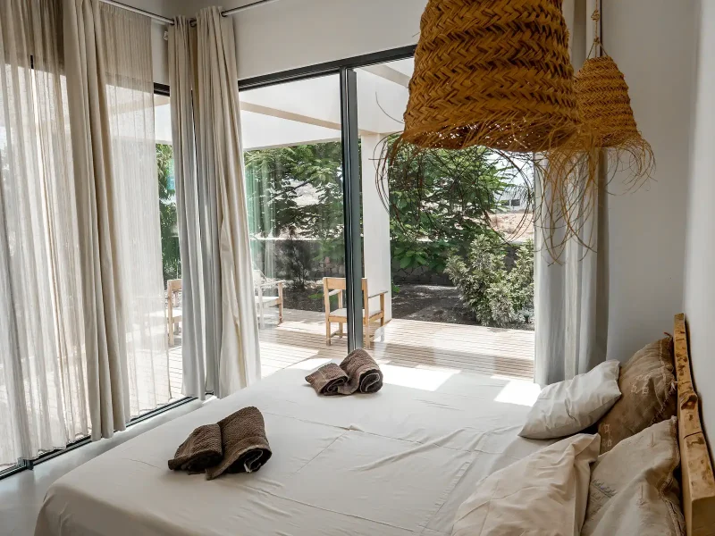 Doppelschlafzimmer im Laola Surf Camp mit weißer Bettwäsche, geflochtener Hängelampe und Glasschiebetüren mit Blick auf private Garten-Terrasse mit Holzdeck.
