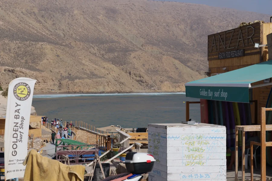 Golden Bay surf shop and Imsouane beachfront with groups on sandy beach and wooden walkway overlooking bay.