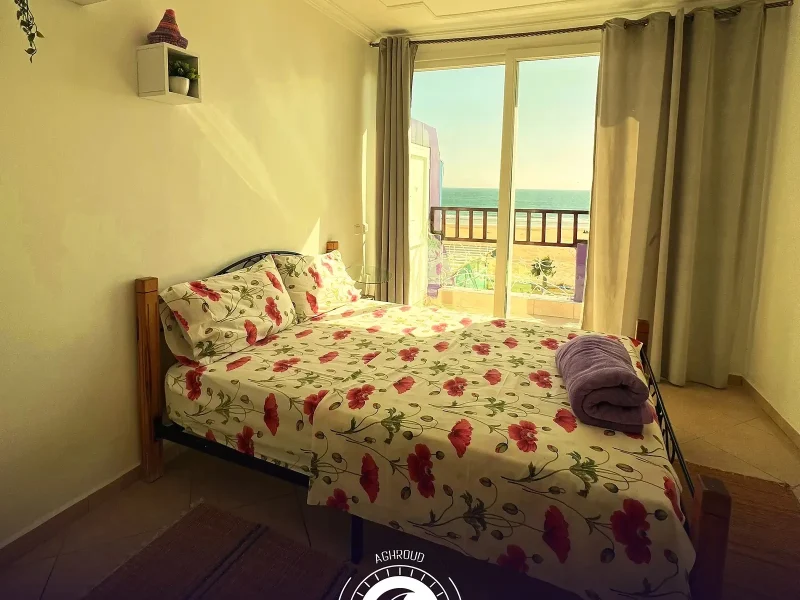 Dormitorio del dúplex en Purple House Surf Camp en Taghazout con vistas al mar a través de las puertas acristaladas y barandilla del balcón visible.