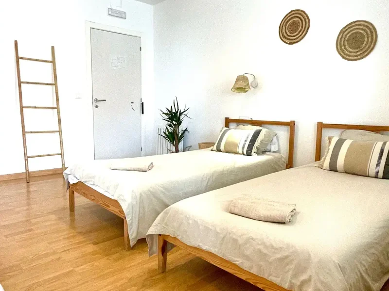 Ein Gemeinschafts-Dreibettzimmer im Laola Galicia mit zwei Holz-Einzelbetten mit cremefarbenen und gestreiften Kissen, weißer Bettwäsche, dekorativen geflochtenen Wandtellern und Holzleiter-Akzent.