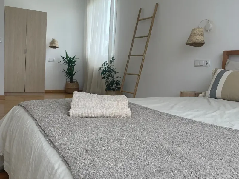Queen-Zimmer im Laola Galicia mit gemachtem Bett mit grauer Tagesdecke und weißen Kissen, Holzkleiderschrank, Topfpflanzen und geflochtenen Hängelampen.