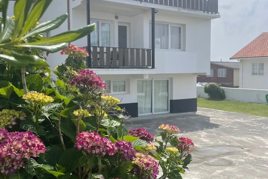 Zweistöckiges weißes Haus im Laola Galicia mit rosa und gelb blühenden Pflanzen, Steinpatio und Balkonen.