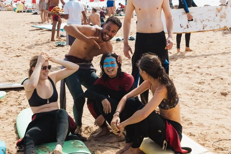 Groupe d'élèves et de moniteurs de surf réunis sur la plage de sable de Taghazout après une session de coaching, avec peinture faciale bleue visible sur un participant.