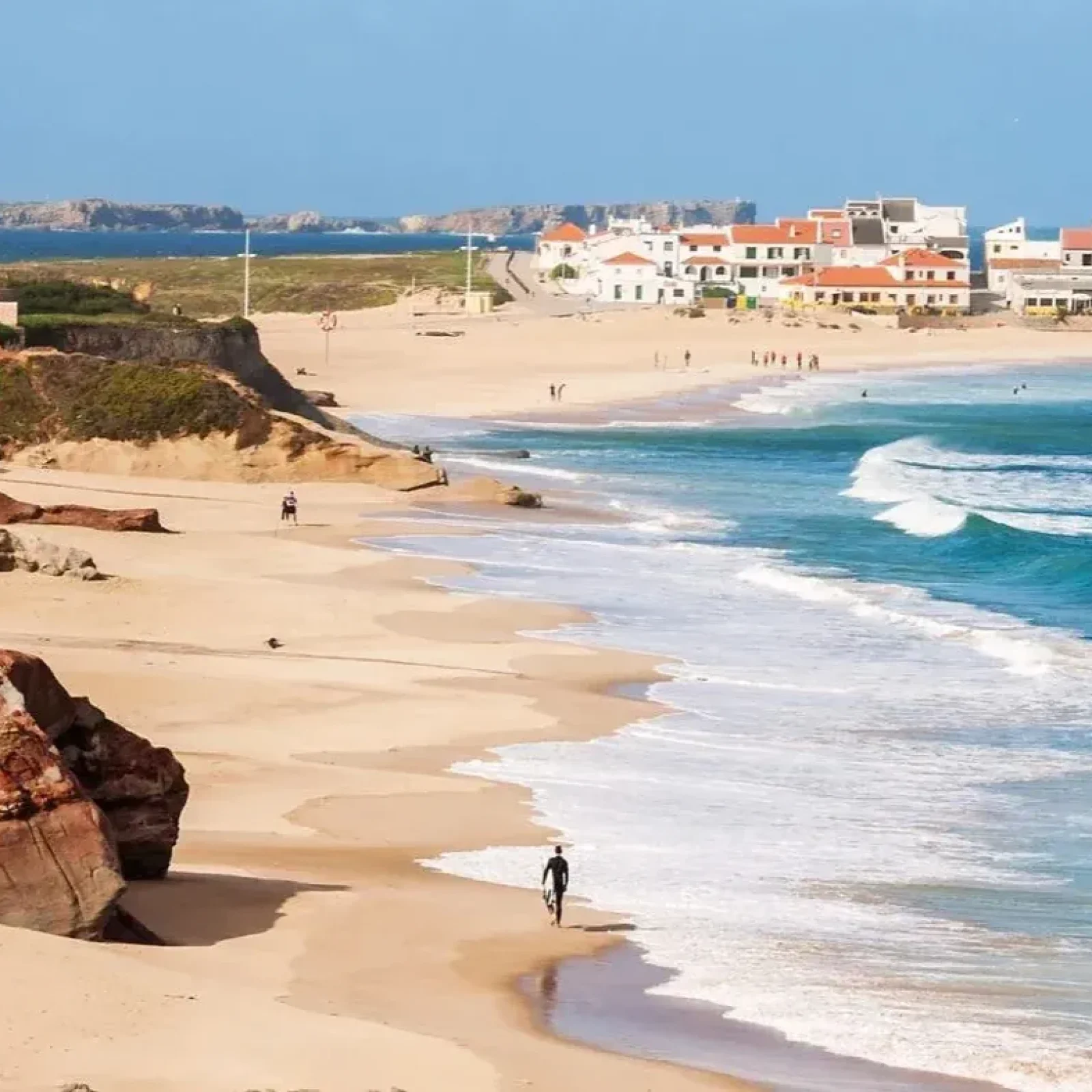 Plage côtière portugaise avec rivages sablonneux, vagues blanches, formations de falaises et bâtiments à toits orange sur le promontoire.