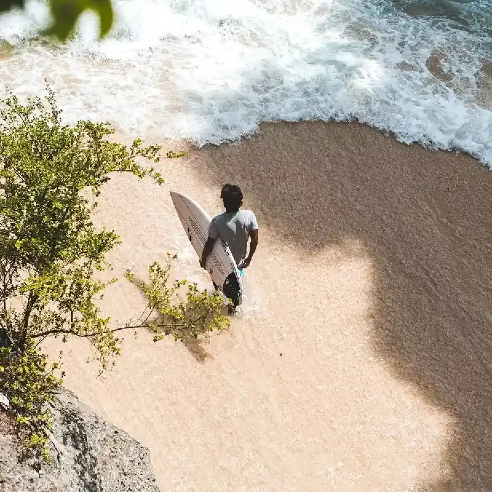 Un surfeur en chemise blanche porte une planche de surf à la plage d'Ahangama avec vagues déferlantes et végétation de mangrove visibles.