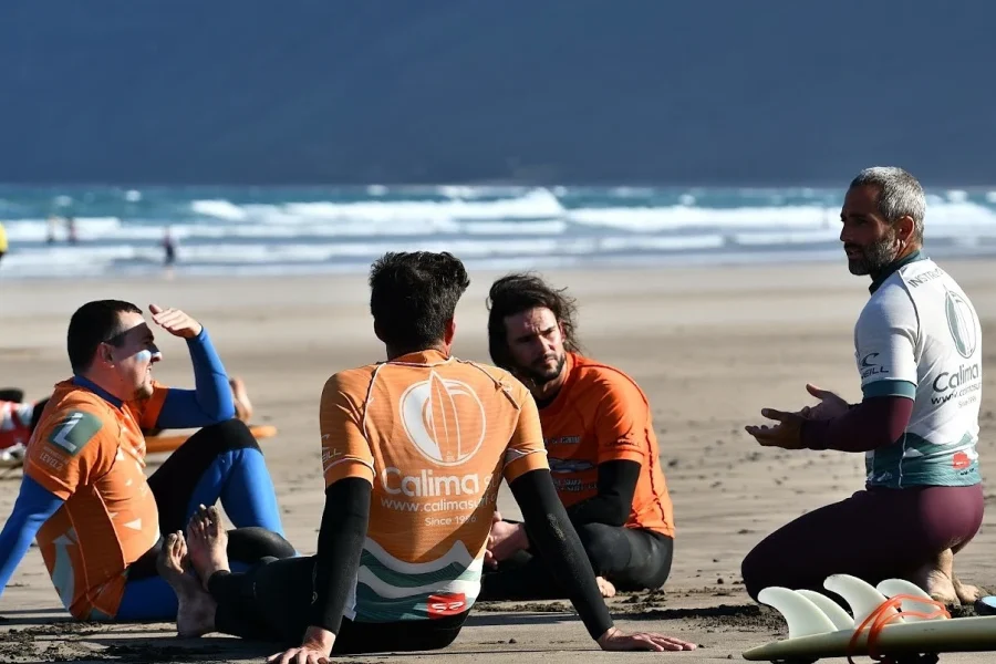 Surflehrer in Neoprenanzügen unterrichten Schüler am Strand von Famara mit rollenden Wellen im Hintergrund.