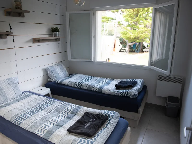 A bright bedroom with two single beds featuring blue geometric patterned bedding, white shiplap walls, and glass sliding doors opening onto a terrace with trees.