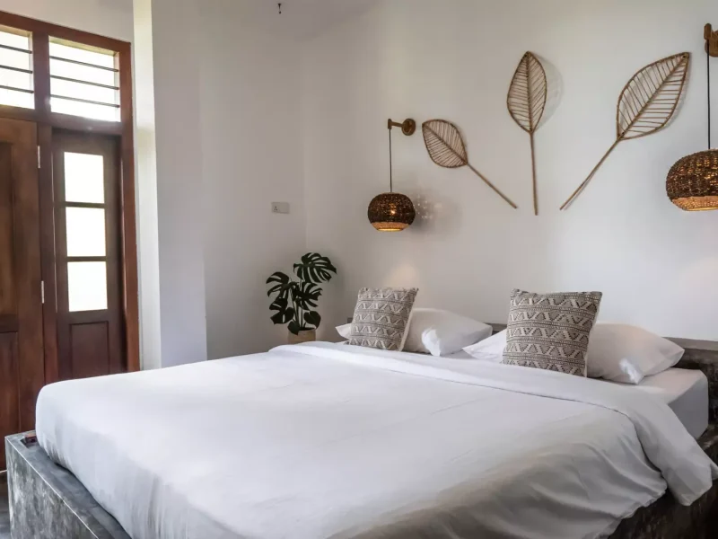 Habitación estándar en Hiriketiya Beach con cama con ropa blanca, cojines estampados, esculturas de hojas tejidas en la pared y lámpara colgante de latón.