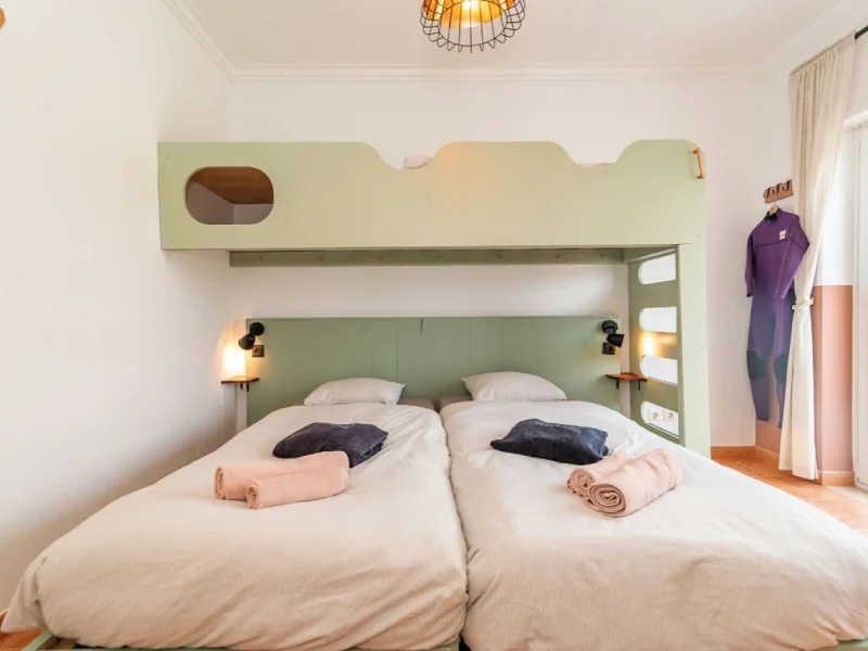 Twin beds with white linens and green padded headboard in the Valley 3 Room at SURFinn Figueira da Foz, featuring rolled towels and blue pillows.