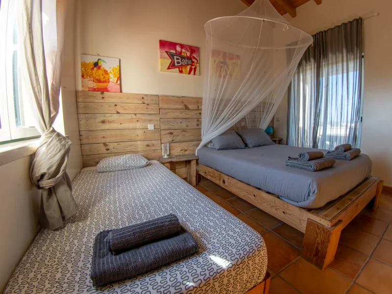 Das Amoreira-Deluxe-Zimmer im Mayla Surf House hat zwei Einzelbetten auf hölzerner Plattform mit weißem Moskitonetz-Baldachin.