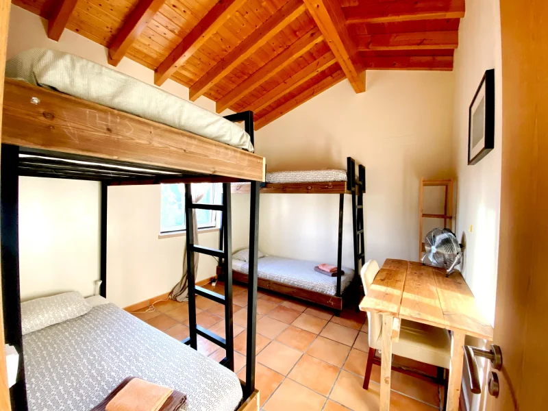 Gemeinschaftszimmer im Mayla Surf House mit zwei Etagenbetten mit schwarzen Metallrahmen, Terrakotta-Fliesenboden und sichtbarer Holzbalkendecke.