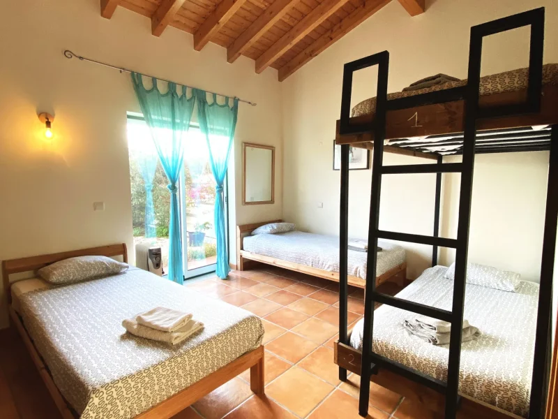 Gemeinschaftszimmer im Mayla Surf House mit Holzbalkendecke, Einzelbett, Etagenbetten und Terrassentür mit türkisfarbenen Vorhängen.