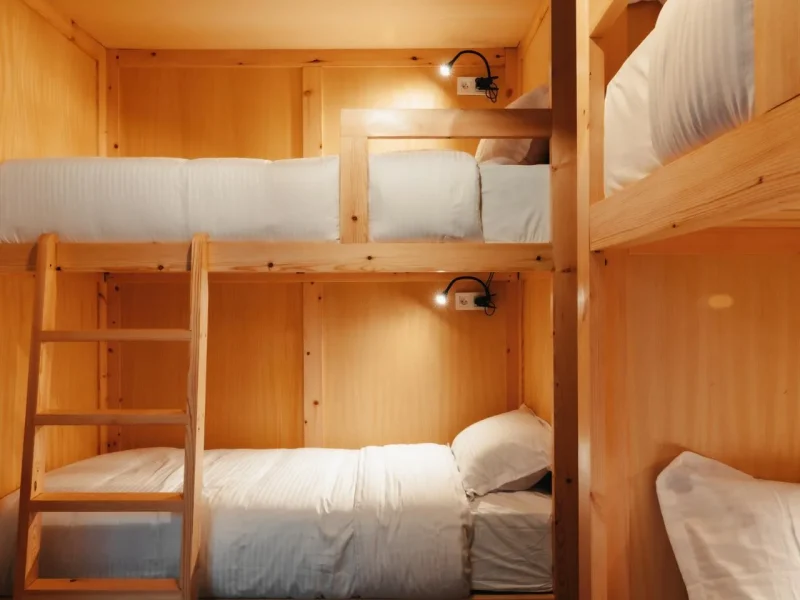 Multi-level wooden bunk bed arrangement with white bedding and individual reading lights in shared dormitory room.