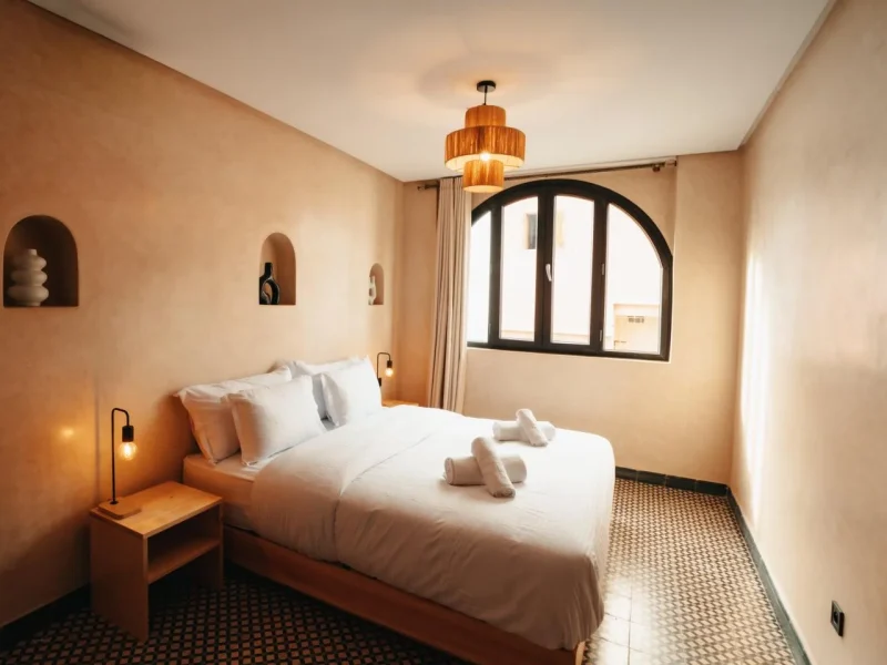 Private double bedroom with white linens, wooden pendant light, arched window with black frame, and neutral beige walls at Gota Dagua Tamraght.
