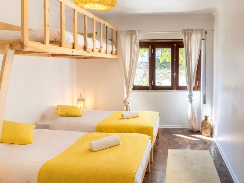 Twin beds with yellow throws in a bright bedroom featuring wooden loft bed, cream curtains, and dark wooden windows at Gota Dagua Surf Camp Caparica.