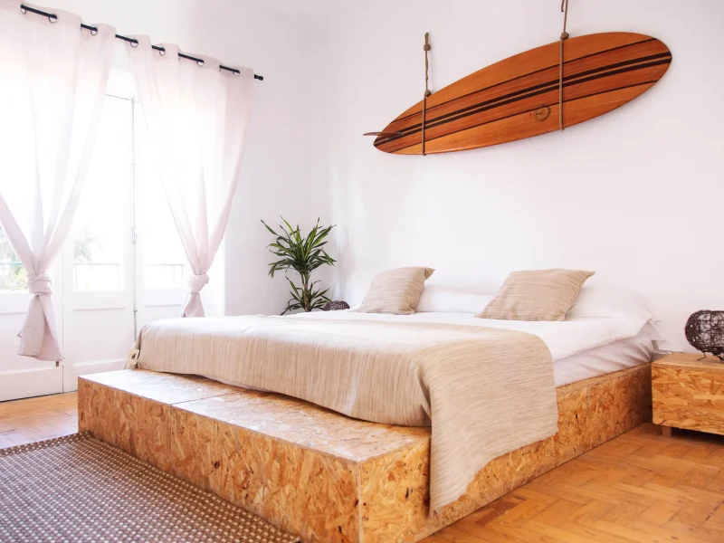 Standard private room at Gota Dagua Surf Camp featuring a cork bed frame with cream bedding, wooden surfboard wall hanging, and pale pink curtains.