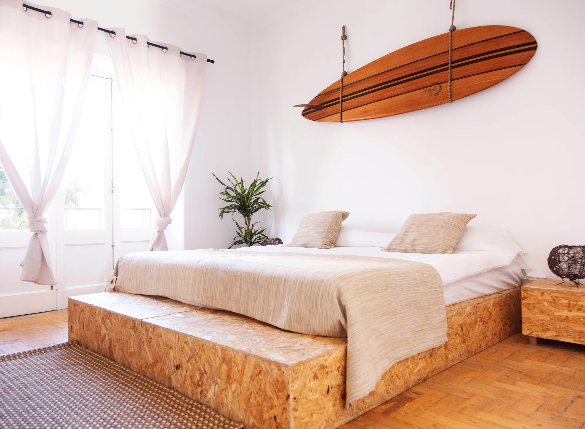 Standard-Privatzimmer im Gota Dagua Surf Camp mit Korkbettrahmen, cremefarbener Bettwäsche, Holzboden und natürlichem Licht aus einem Fenster mit Blick auf die Caparica-Küste.