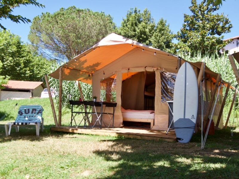 Family Safaritent im Familycamp Zarautz im Baskenland mit offener Terrasse, orangefarbenem Stoffdach und Camping-Einrichtung inklusive Schreibtisch und Surfbrett.
