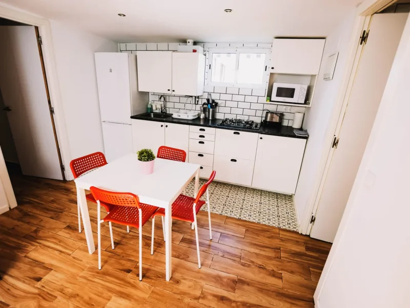 Une cuisine lumineuse dans un bungalow présente un mobilier blanc, plan de travail noir, crédence en carrelage métro et table à manger avec chaises rouges.