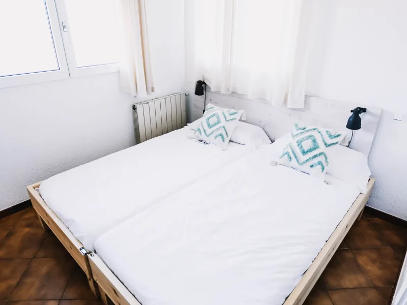 Chambre Double Deluxe à Surfbase Loredo présentant un lit à cadre en bois avec linge blanc et oreillers géométriques turquoise, murs blancs et sol en terracotta.