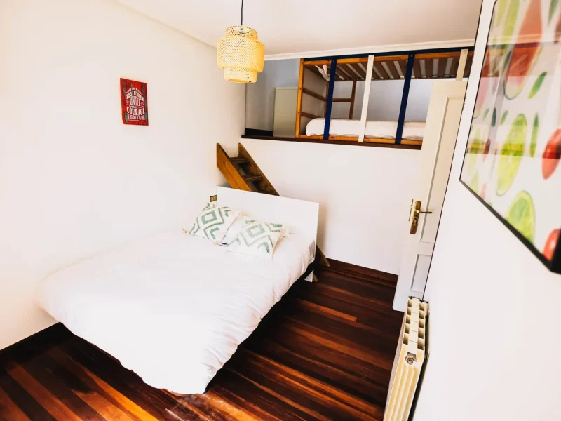 Chambre Four Standard à Surfbase Loredo présentant un lit simple avec linge blanc et oreillers à motifs, avec lit superposé en mezzanine au-dessus, suspension jaune et murs blancs.
