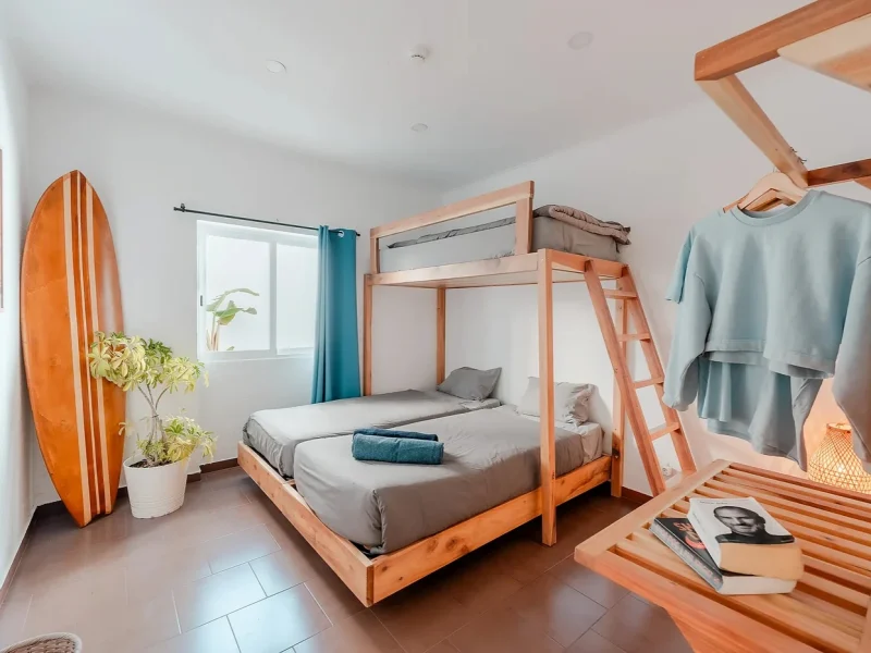 Shared bedroom with wooden bunk bed frame, gray bedding, teal curtains, and potted plant by window in Lisbon.