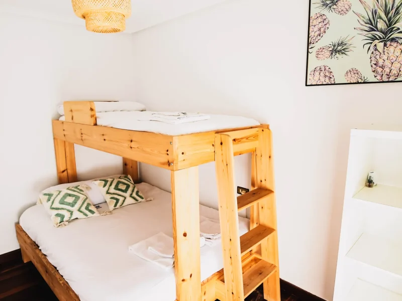 Intérieur de chambre Triple Standard avec lit superposé en bois, literie blanche, œuvre murale d'ananas et lampe tissée au plafond.