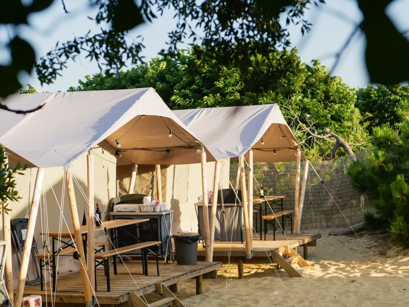 Multiple canvas bell tents with wooden platforms situated on a sandy beach with green vegetation visible in background.