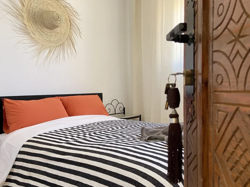 Double room interior with black and white striped blanket, orange pillows, wooden door and straw wall decoration.