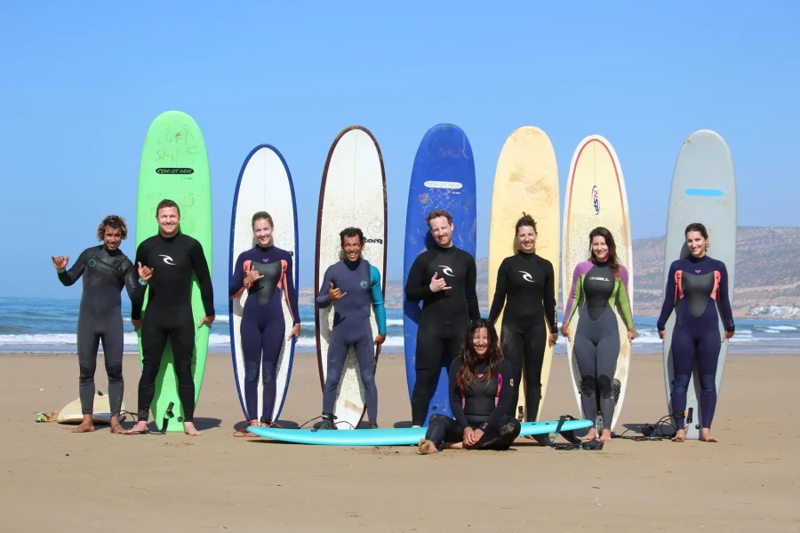 Seven surfers holding multiple colorful surfboards on a sandy beach with ocean and hills visible in background at Taghazout.