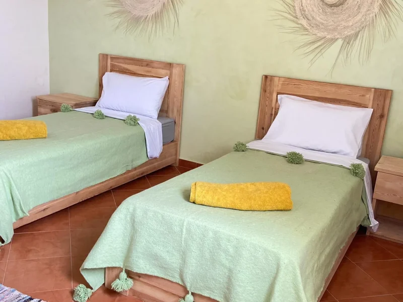 Twin bedroom at Shaka Surf Morocco features two wooden-framed beds with green coverlets and decorative woven wall hangings.
