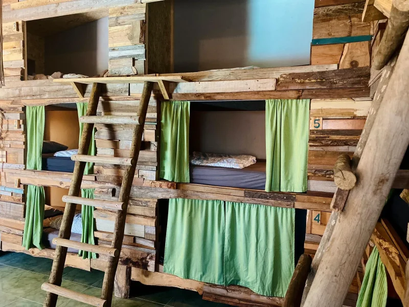 9 bed pod dorm room at Stoke Surf House with wooden bunk structures and green fabric privacy curtains.