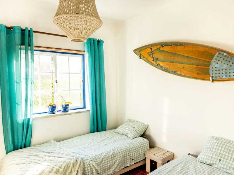 Double bedroom at Stoke Surf House featuring twin beds with patterned bedding, blue-framed window with turquoise curtains, woven pendant lamp, and vintage surfboard mounted on wall.