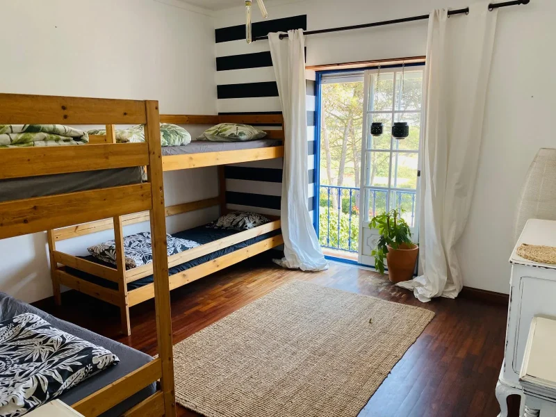 4-bed female dormitory with wooden bunk beds, black and white striped accent wall, and glass doors opening to a balcony overlooking trees.