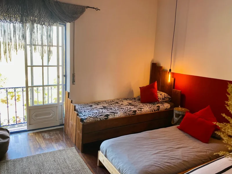 Private triple ensuite bedroom at Stoke Surf House Portugal featuring two single beds with patterned bedding and red accent pillows.