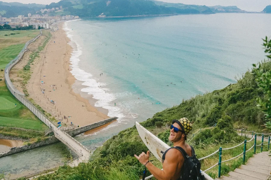 Surfer holding board on clifftop stairs overlooking Ondarreta Beach and the Basque coast with residential areas and forested headland.