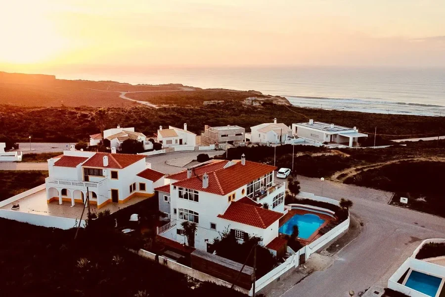 Vista aérea del resort SURFinn Algarve con villas blancas, tejados de terracota, piscina azul y acantilados costeros con vistas al océano Atlántico al atardecer.