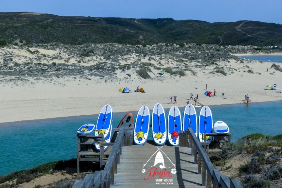 Row of blue and white SUP paddleboards moored at wooden dock in Vale da Telha with sandy beach, dunes and beach visitors visible in background.