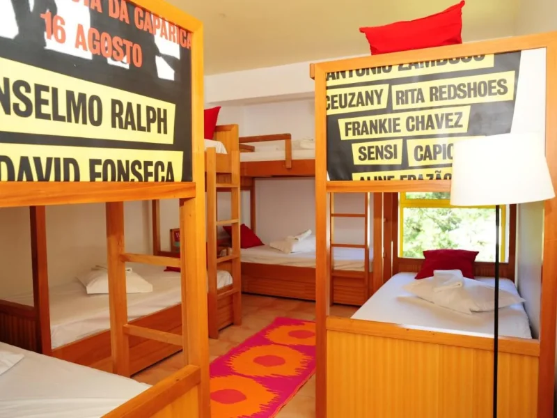 Large shared dorm room with multiple wooden bunk beds, red and orange bedding, and signage listing guest names on the walls at Gota Dagua Junior Surf Camp in Caparica.