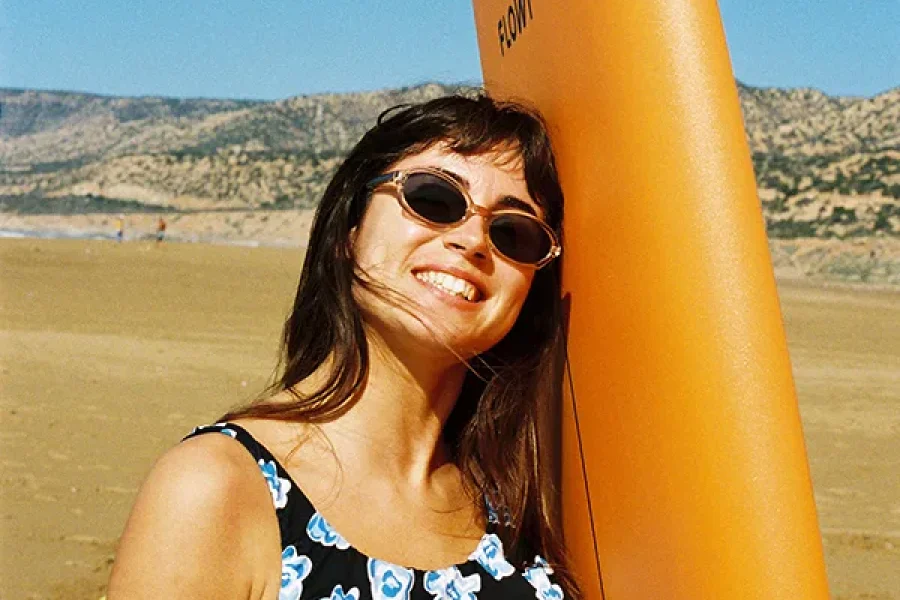 Woman with surfboard at Tamraght beach, Morocco with coastal hills visible in background.